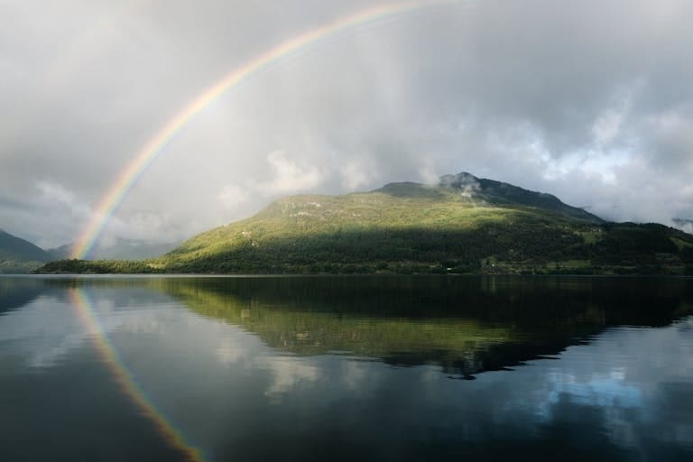 A stunning rainbow arches over a peaceful fjord in Sandeid, Norway, reflecting its beauty on the tranquil water surface.